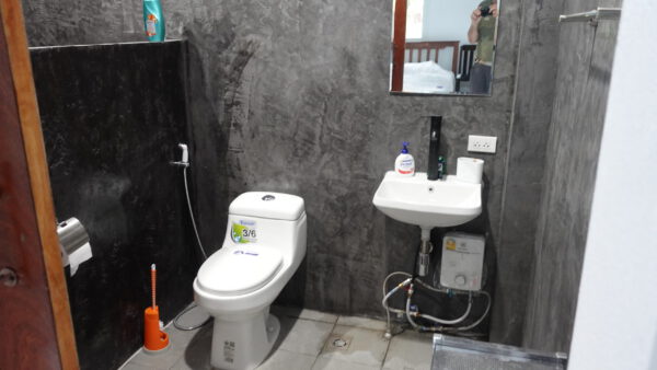 Das Bad mit europäischem WC, Waschbecken und Spiegel vor dunkel gespachtelten Betonwänden; rechts an der Wand hängt der Durchlauferhitzer.