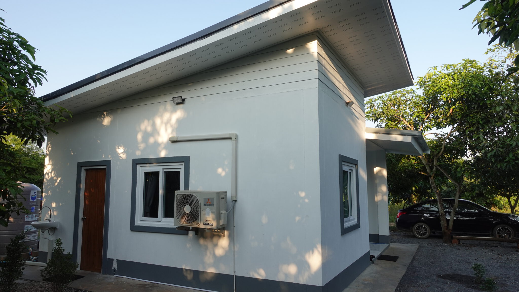 Das Haus von der Ecke im tiefstehenden Abendlicht: ein weit auskragendes Pultdach, das Außengerät der Klimaanlage an der Wand, links daneben der Wassertank.