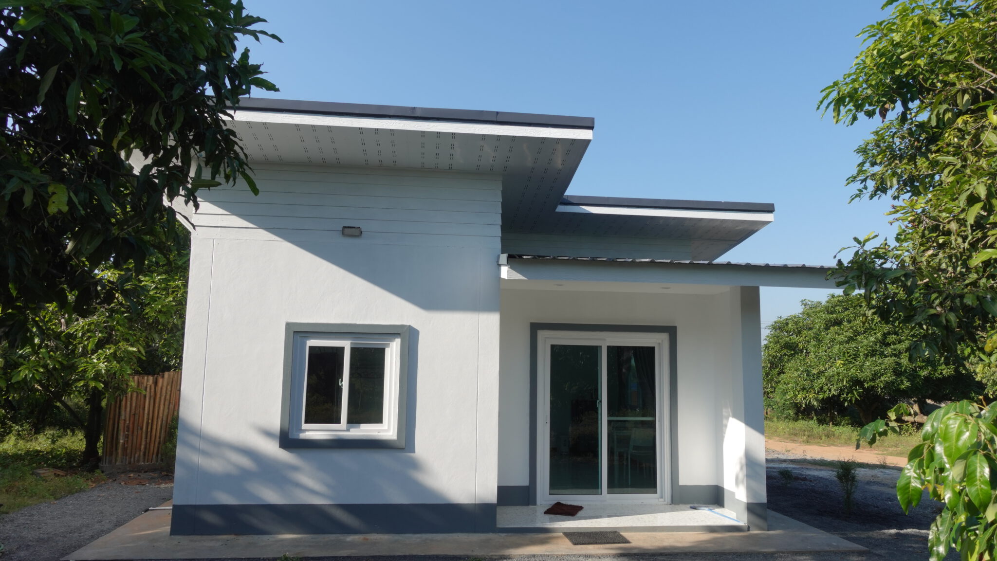 Das Gästehaus von vorn: ein weißer Flachdachbau mit grau abgesetzten Fensterrahmen, eine große Glasschiebetür führt auf die überdachte Terrasse, ringsum Mangobäume.