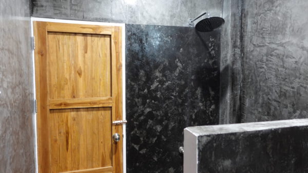 Die Dusche im Bad: ein Regenduschkopf über einer gemauerten Abtrennung, dunkel gespachtelte Betonwände, daneben eine Tür aus hellem Holz.