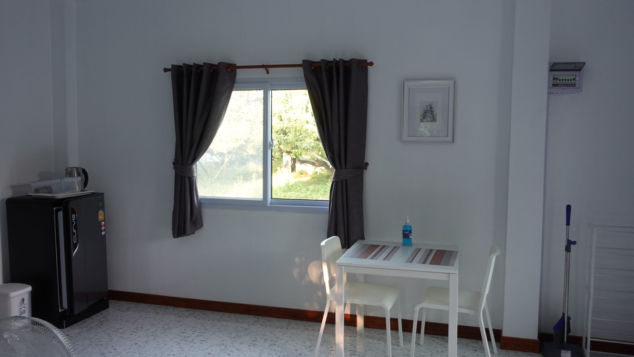 Der Wohnbereich: ein weißer Esstisch mit zwei Stühlen unter dem Fenster, links ein Kühlschrank mit Wasserkocher darauf, heller Terrazzoboden.